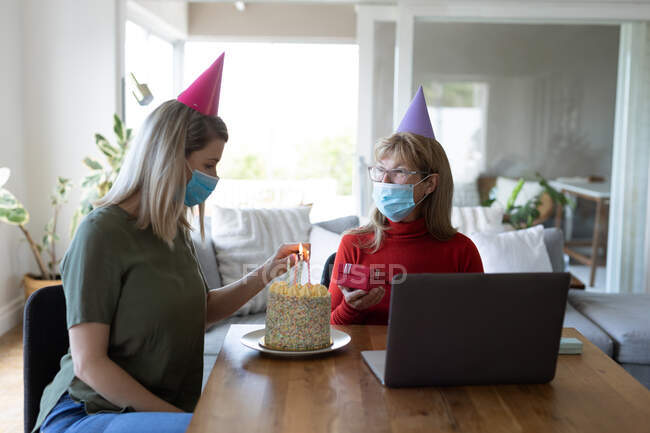 Senior Caucasian woman spending time at home with her adult daughter, sitting in living room with a birthday cake and using laptop. Social distancing during Covid 19 Coronavirus quarantine. — Stock Photo