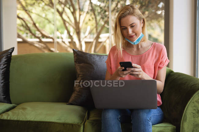 Mulher branca trabalhando em casa, usando máscara facial, usando smartphone e laptop. Distanciamento social durante o bloqueio de quarentena do Covid 19 Coronavirus. — Fotografia de Stock