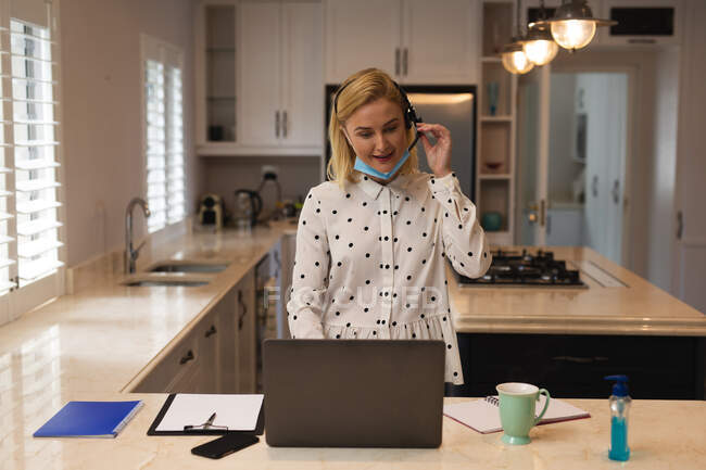 Mulher caucasiana trabalhando em casa, usando máscara facial, usando fone de ouvido e computador portátil. Distanciamento social durante o bloqueio de quarentena do Covid 19 Coronavirus. — Fotografia de Stock