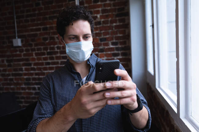 Kaukasischer Mann, der in einem lässigen Büro arbeitet, sein Smartphone benutzt und Gesichtsmaske trägt. Soziale Distanzierung am Arbeitsplatz während der Coronavirus Covid 19 Pandemie. — Stockfoto