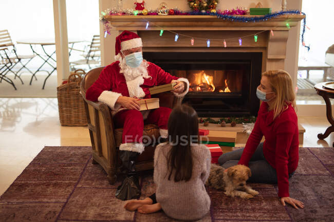 Senior Caucasian man, his adult daughter and granddaughter at home dressed as Father Christmas, wearing face mask, giving presents. Social distancing during Covid 19 Coronavirus quarantine lockdown. — Stock Photo
