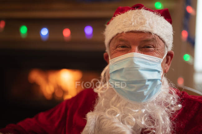Ritratto di anziano caucasico a casa vestito da Babbo Natale, mascherato, che guarda la macchina fotografica. Distanza sociale durante il blocco di quarantena Covid 19 Coronavirus. — Foto stock
