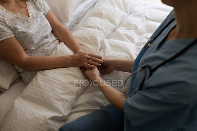 Frau zu Hause von einer Krankenschwester besucht, auf dem Bett sitzend, Händchen haltend. Medizinische Versorgung zu Hause während der Quarantäne des Covid 19 Coronavirus. — Stockfoto