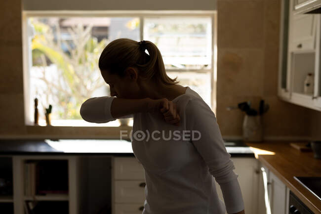 Kaukasierin, die in der Küche steht und in ihren Ellbogen hustet. Medizinische Versorgung zu Hause während der Quarantäne des Covid 19 Coronavirus. — Stockfoto