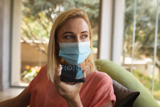 Mujer caucásica pasando tiempo en casa, usando mascarilla, hablando en un teléfono inteligente. Distanciamiento social durante el bloqueo de cuarentena del Coronavirus Covid 19. - foto de stock
