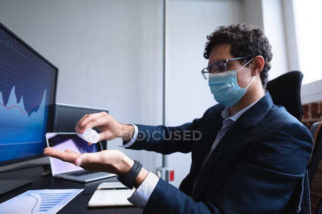 Hombre caucásico trabajando en una oficina informal, usando desinfectante y usando mascarilla facial. Distanciamiento social en el lugar de trabajo durante la pandemia de Coronavirus Covid 19. - foto de stock