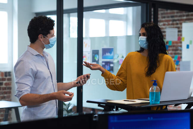 Смешанная расовая женщина и белый мужчина работают в офисе, носят маски и разговаривают. Социальное дистанцирование на рабочем месте во время пандемии Coronavirus Covid 19. — стоковое фото