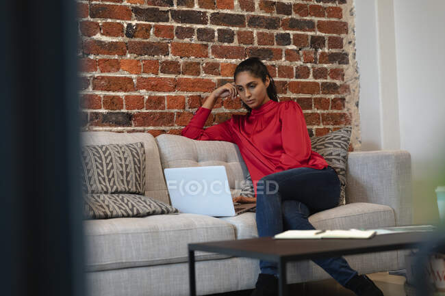 Donna di razza mista che lavora in un ufficio informale, seduta su un divano, usando un computer portatile. Distanze sociali sul luogo di lavoro durante la pandemia di Coronavirus Covid 19. — Foto stock