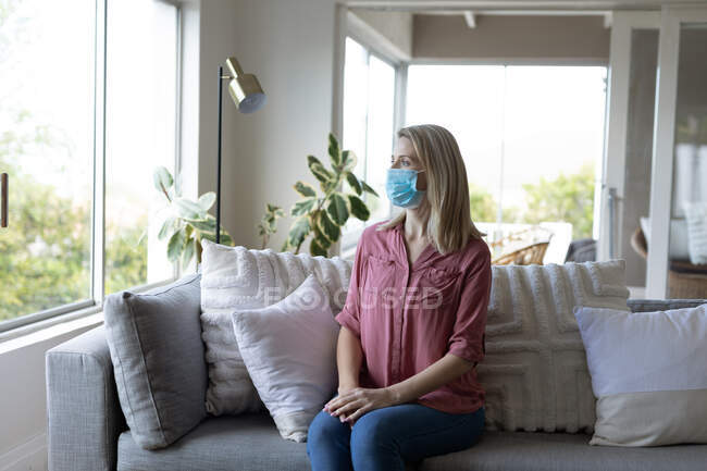 Mujer caucásica pasando tiempo en casa, sentada en un sofá, con una máscara facial. Distanciamiento social durante la cuarentena del Coronavirus Covid 19. - foto de stock