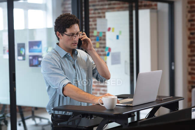 Homme caucasien travaillant dans un bureau décontracté, parlant sur un smartphone et utilisant un ordinateur portable. Distance sociale sur le lieu de travail pendant la pandémie de coronavirus Covid 19. — Photo de stock