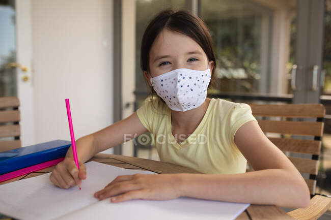 Retrato de una chica caucásica pasando tiempo en casa, usando mascarilla, haciendo tareas escolares. Distanciamiento social durante el bloqueo de cuarentena del Coronavirus Covid 19. - foto de stock