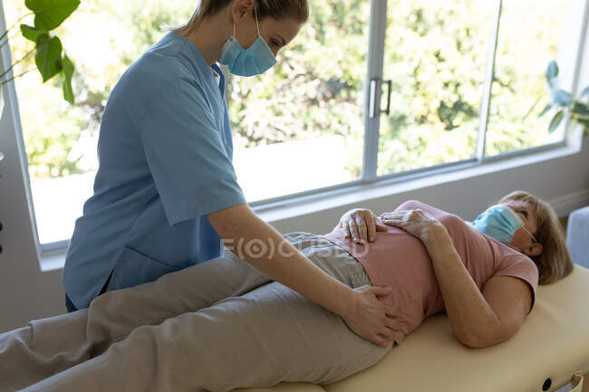 Mujer mayor caucásica en casa visitada por una enfermera caucásica, estirando sus caderas, usando máscaras faciales. Atención médica en el hogar durante la cuarentena del Coronavirus de Covid 19. - foto de stock
