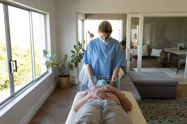 Mulher branca sênior em casa visitada por enfermeira caucasiana, massageando o pescoço, usando máscaras faciais. Carro médico — Fotografia de Stock