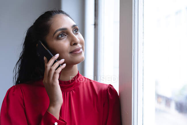 Mujer de raza mixta trabajando en una oficina informal, hablando en un teléfono inteligente. Distanciamiento social en el lugar de trabajo durante la pandemia de Coronavirus Covid 19. - foto de stock