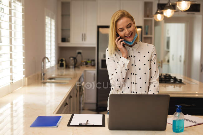 Белая женщина работает из дома, носит маску, разговаривает на смартфоне и использует ноутбук. Социальное дистанцирование во время изоляции коронавируса Covid 19. — стоковое фото