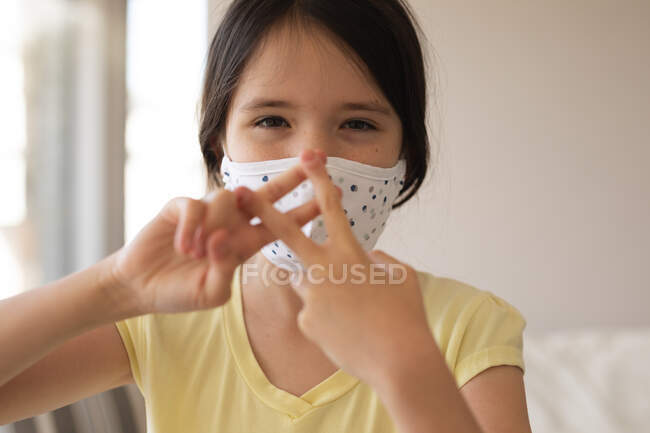 Retrato de menina caucasiana passar o tempo em casa, usando máscara facial, olhando para a câmera, usando linguagem gestual. Distanciamento social durante o bloqueio de quarentena do Covid 19 Coronavirus. — Fotografia de Stock