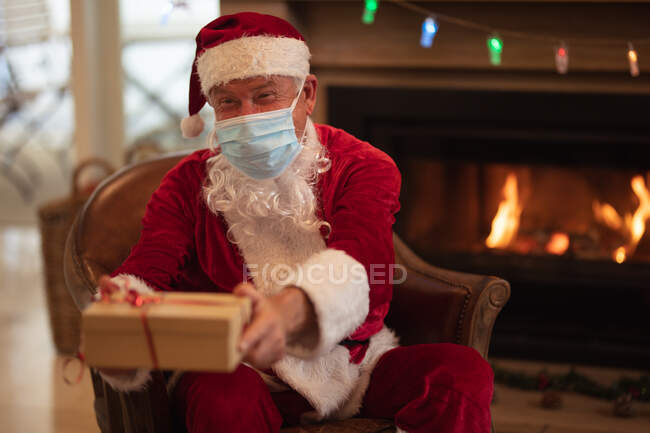 Ein älterer kaukasischer Mann zu Hause, als Weihnachtsmann verkleidet, mit Gesichtsmaske, sitzt auf einem Stuhl am Kamin und verteilt Geschenke. Soziale Distanzierung während Covid 19 Coronavirus Quarantäne Lockdown. — Stockfoto