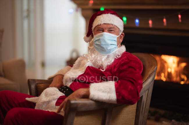 Homem caucasiano sênior em casa vestido como Pai Natal, usando máscara facial, sentado em uma cadeira à lareira. Distanciamento social durante o bloqueio de quarentena do Covid 19 Coronavirus. — Fotografia de Stock