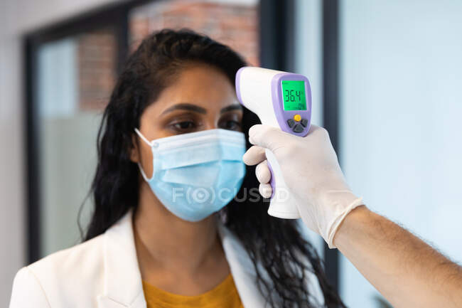 Mixed race woman and Caucasian man working in a casual office, wearing face mask, a man taking her temperature. Social distancing in the workplace during Coronavirus Covid 19 pandemic. — Stock Photo