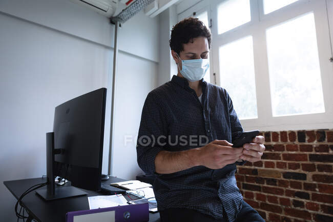 Кавказький чоловік працює в повсякденному офісі, використовуючи свій смартфон і маску обличчя. Суспільна дистанція на робочому місці під час Коронавірусу Ковід 19 пандемії. — стокове фото