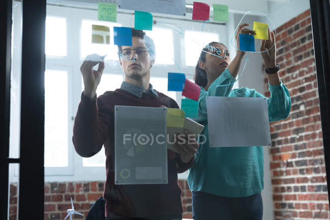 Смешанная расовая женщина и белый мужчина работают в офисе, пишут на стеклянной доске и разговаривают. Креативные профессионалы бизнеса, работающие в современном офисе. — стоковое фото