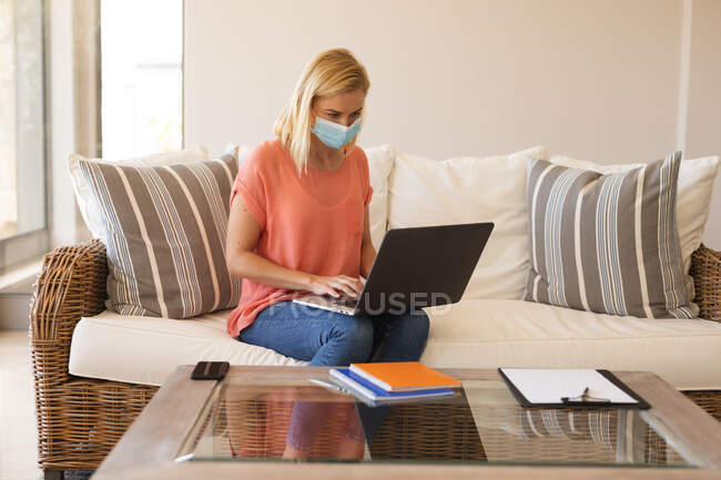 Kaukasische Frau, die von zu Hause aus arbeitet, Gesichtsmaske trägt und Laptop benutzt. Soziale Distanzierung während Covid 19 Coronavirus Quarantäne Lockdown. — Stockfoto