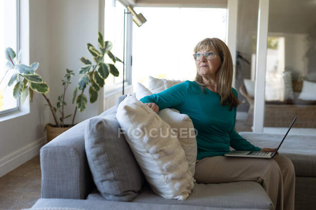 Mujer mayor caucásica pasar tiempo en casa, sentado en su sala de estar con una computadora portátil. Distanciamiento social durante el bloqueo de cuarentena del Coronavirus Covid 19. - foto de stock