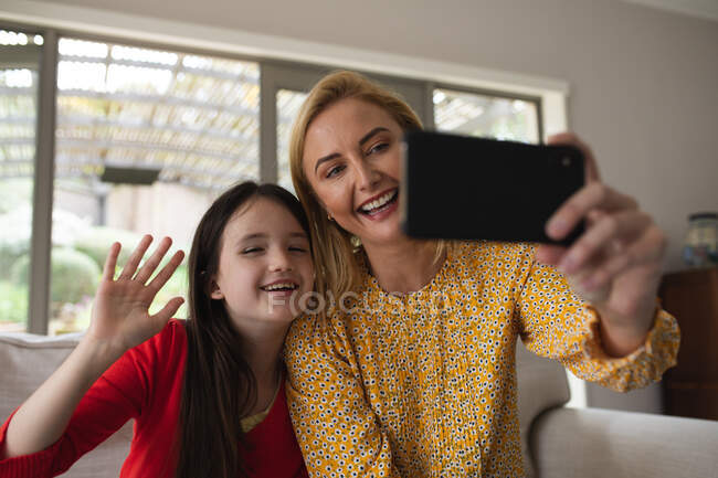 Caucasian woman and her daughter spending time at home together, using smartphone, making a video call. Social distancing during Covid 19 Coronavirus quarantine lockdown. — Stock Photo