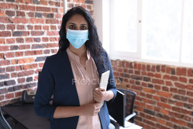 Portrait of mixed race woman working in a casual office, holding tablet, wearing face mask and looking at camera. Social distancing in the workplace during Coronavirus Covid 19 pandemic. — Stock Photo