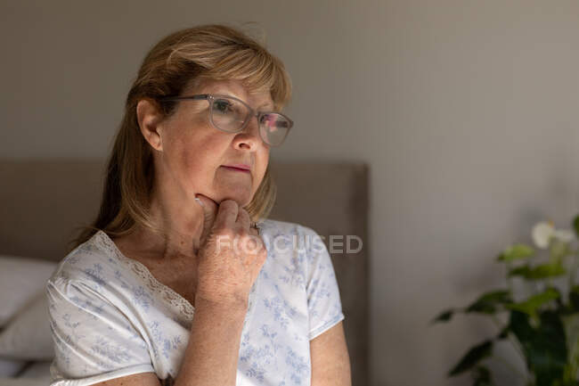 Старшая белая женщина проводит время дома, сидит в своей спальне, смотрит в сторону и думает. Социальное дистанцирование во время изоляции коронавируса Covid 19. — стоковое фото