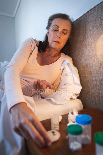 Больная белая женщина проводит время дома, социальное дистанцирование и самоизоляция в карантинной изоляции, лежит в постели, держит таблетки. — стоковое фото