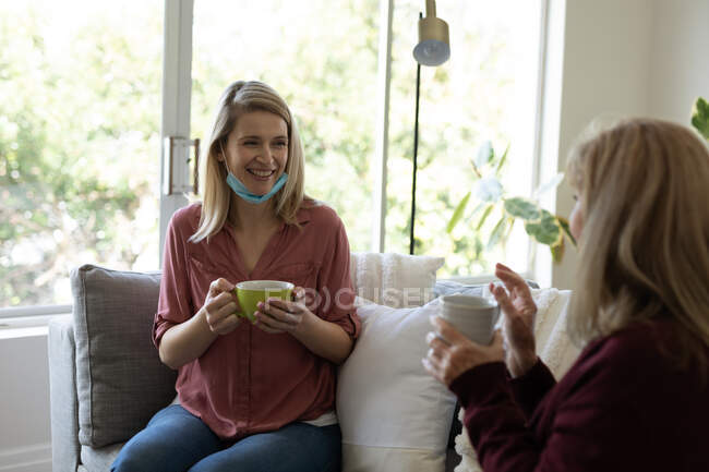 Senior Caucasian woman spending time at home with her adult daughter, sitting on couch, drinking cup of tea. Social distancing during Covid 19 Coronavirus quarantine. — Stock Photo