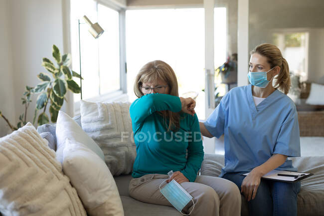 Mujer caucásica siendo visitada en casa por una enfermera caucásica, cubriendo la boca mientras tose, la enfermera con mascarilla facial. Atención médica en el hogar durante la cuarentena del coronavirus de Covid 19. - foto de stock