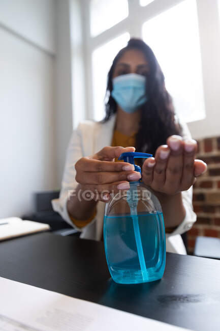 Close up of mixed race woman working in a casual office, using sanitizer and wearing face mask. Social distancing in the workplace during Coronavirus Covid 19 pandemic. — Stock Photo