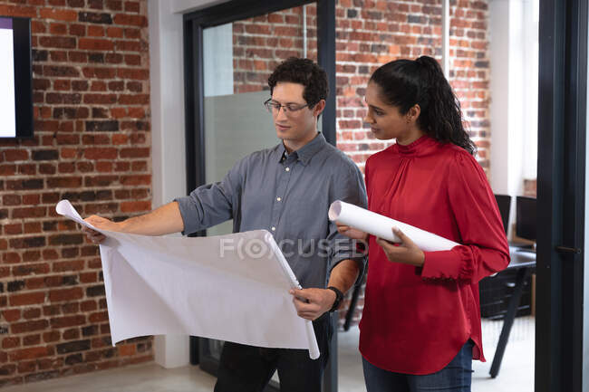 Смешанная расовая женщина и кавказский мужчина работают в случайном офисе, держат планы и разговаривают. Креативные профессионалы бизнеса, работающие в современном офисе. — стоковое фото