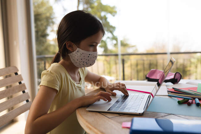 Kaukasisches Mädchen, das Zeit zu Hause verbringt, Gesichtsmaske trägt und während des Online-Schulunterrichts einen Laptop benutzt. Soziale Distanzierung während Covid 19 Coronavirus Quarantäne Lockdown. — Stockfoto