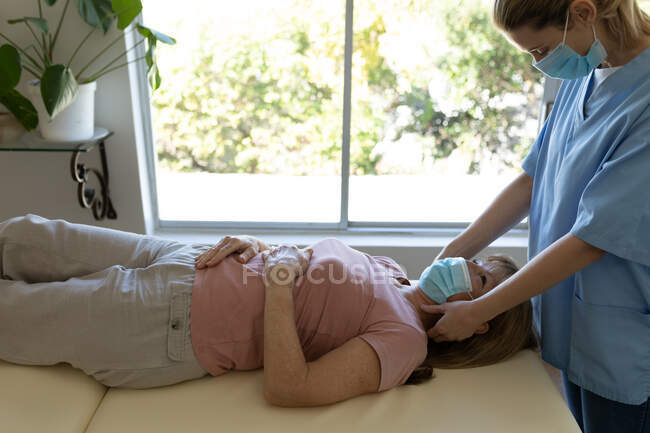 Senior Caucasian woman at home visited by Caucasian female nurse, massaging her neck, wearing face masks. Medical car — Stock Photo