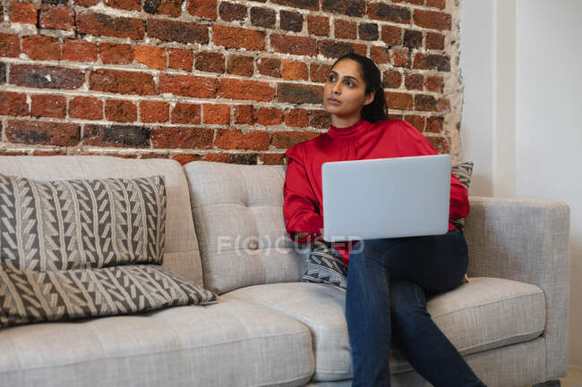 Змішана расова жінка працює в повсякденному офісі, сидячи на дивані, за допомогою ноутбука. Суспільна дистанція на робочому місці під час Коронавірусу Ковід 19 пандемії. — стокове фото