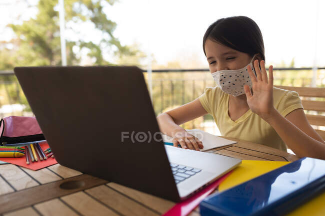 Une fille caucasienne passe du temps à la maison, portant un masque facial, utilisant un ordinateur portable pendant la leçon d'école en ligne. Distance sociale pendant le confinement en quarantaine du coronavirus Covid 19. — Photo de stock