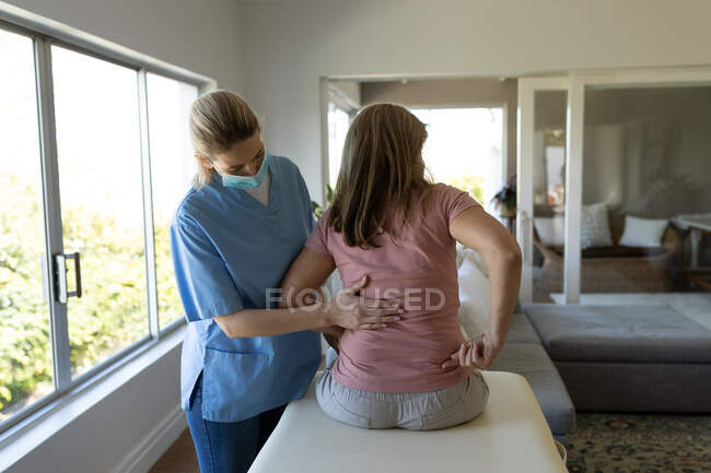 Mujer mayor caucásica en casa visitada por una enfermera caucásica, estirando la espalda, enfermera con máscara facial. Atención médica en el hogar durante la cuarentena del Coronavirus de Covid 19. - foto de stock