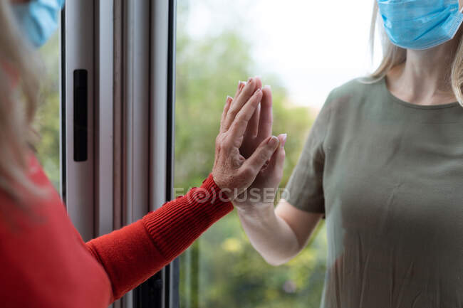 Старшая белая женщина и ее взрослая дочь дома, в масках и приветствуют друг друга прикосновением рук. Социальное дистанцирование, здоровье и гигиена во время пандемии коронавируса Covid 19. — стоковое фото