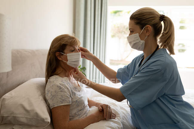 Mulher branca sênior em casa visitada por enfermeira caucasiana, colocando uma máscara facial, enfermeira vestindo máscara facial. — Fotografia de Stock