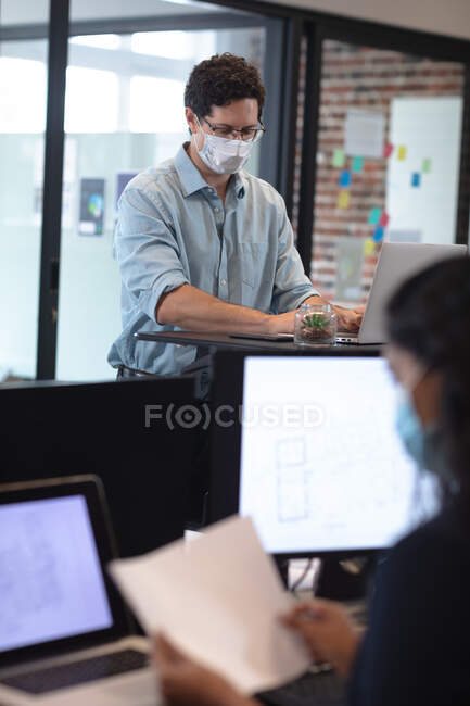 Hombre caucásico y mujer de raza mixta trabajando en una oficina informal, usando máscaras faciales, utilizando computadoras portátiles. Distanciamiento social en el lugar de trabajo durante la pandemia de Coronavirus Covid 19. - foto de stock