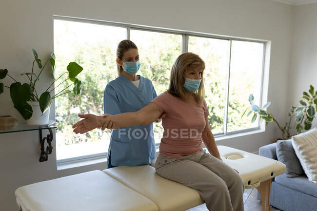 Mujer mayor caucásica en casa visitada por una enfermera caucásica, estirando el brazo, usando máscaras faciales. Atención médica en el hogar durante la cuarentena del Coronavirus de Covid 19. - foto de stock