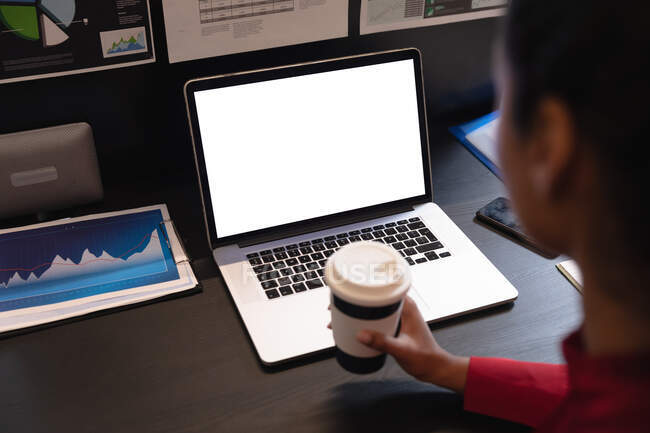 Mulher de raça mista trabalhando em um escritório casual, sentado na mesa, usando um computador portátil, segurando o café takeaway. Distanciamento social no local de trabalho durante a pandemia do Coronavirus Covid 19. — Fotografia de Stock