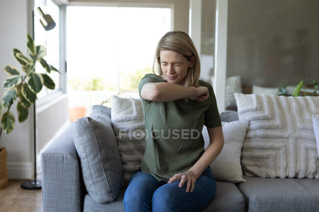 Donna caucasica che passa del tempo a casa, seduta sul divano, starnutisce nel gomito. Distanze sociali durante la quarantena di Covid 19 Coronavirus. — Foto stock