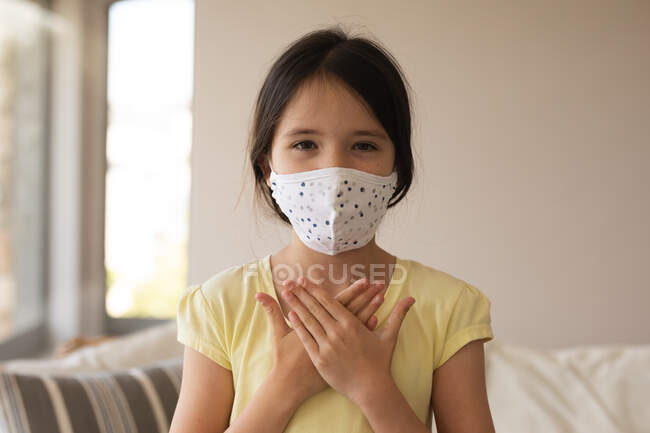 Portrait of Caucasian girl spending time at home, wearing face mask, looking at camera, using sign language. Social distancing during Covid 19 Coronavirus quarantine lockdown. — Stock Photo
