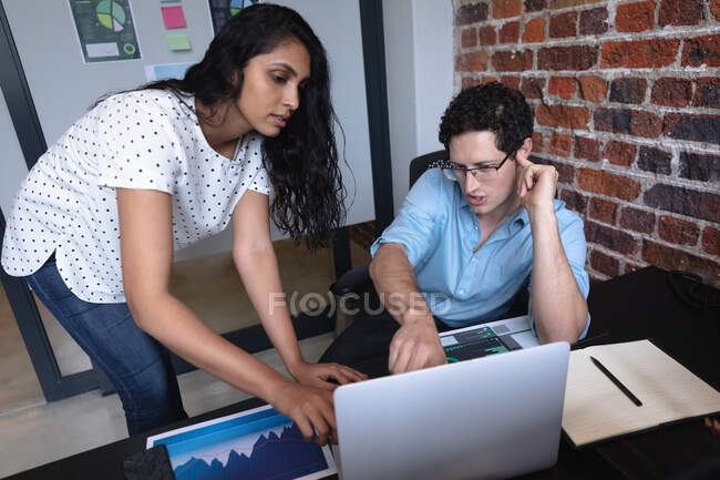 Смешанная расовая женщина и белый мужчина работают в обычном офисе, используют ноутбук и обсуждают свою работу. Креативные профессионалы бизнеса, работающие в современном офисе. — стоковое фото