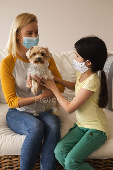Mujer caucásica y su hija pasando tiempo en casa juntos, usando máscaras faciales, jugando con su perro. Distanciamiento social durante el bloqueo de cuarentena del Coronavirus Covid 19. - foto de stock