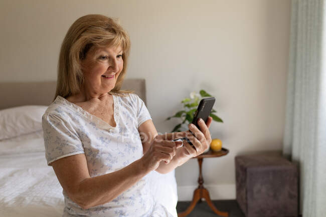 Mujer mayor caucásica pasar tiempo en casa, sentado en su sala de estar con un teléfono inteligente. Distanciamiento social durante el bloqueo de cuarentena del Coronavirus Covid 19. - foto de stock